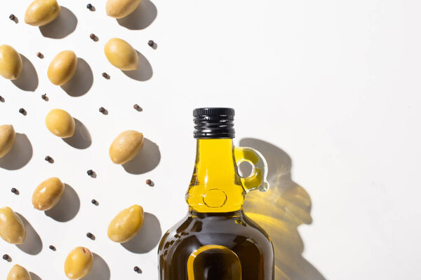 top view of olive oil in bottle near green olives and black pepper on white background with shadow