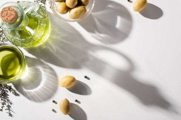 top view of olive oil in bottle and bowl near herb, green olives and black pepper on white background with shadow