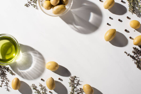 top view of olive oil in bottle near green olives and black pepper in shadow on white background