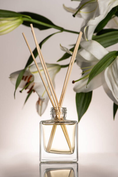 селективный фокус деревянных палочек в духах в бутылке рядом с цветами лилии на бежевом фоне

