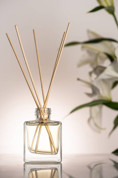 selective focus of wooden sticks in perfume in bottle near lily flowers on beige background