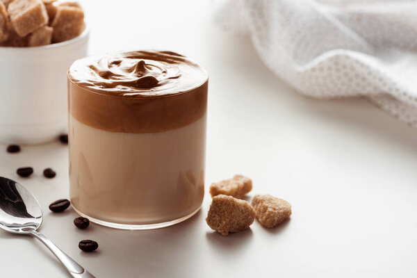 selective focus of delicious Dalgona coffee in glass near napkin, spoon, coffee beans and brown sugar on white background