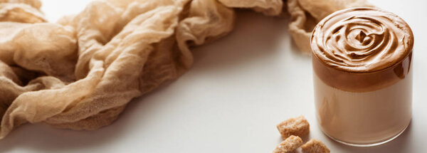 delicious Dalgona coffee in glass near cloth and granulated brown sugar on white background, panoramic shot
