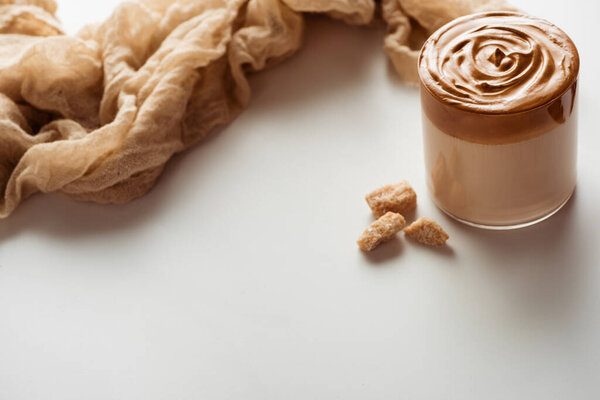 delicious Dalgona coffee in glass near cloth and granulated brown sugar on white background