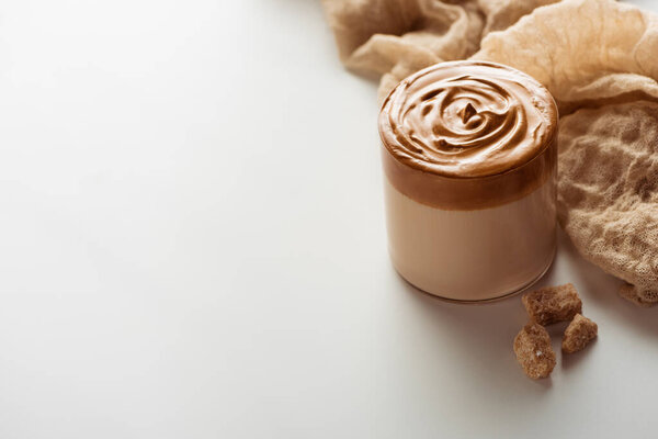 delicious Dalgona coffee in glass near cloth and granulated brown sugar on white background