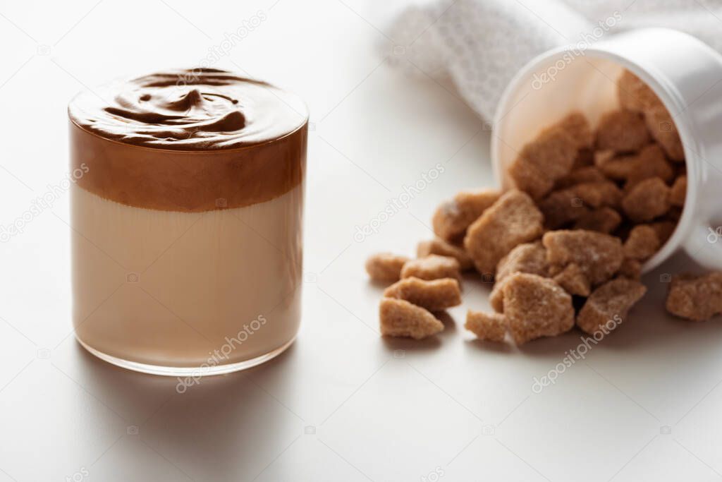 selective focus of delicious Dalgona coffee in glass near scattered brown sugar on white background