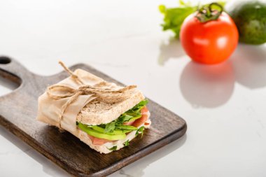 selective focus of fresh green sandwich with avocado and prosciutto on wooden cutting board on white marble surface clipart