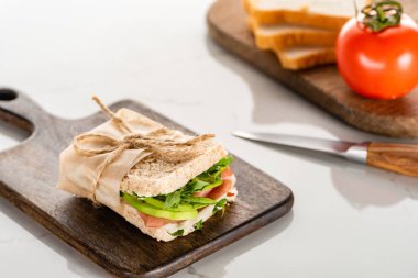 selective focus of fresh green sandwich with avocado and prosciutto on wooden cutting board on white marble surface clipart