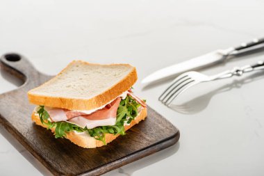 Beyaz mermer yüzeydeki çatal bıçak takımının yanında roka ve prosciutto ile taze sandviçin seçici odak noktası.