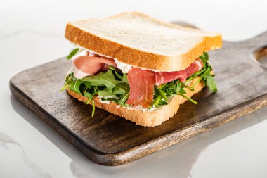 selective focus of fresh green sandwich with arugula and prosciutto on wooden cutting board on white marble surface clipart