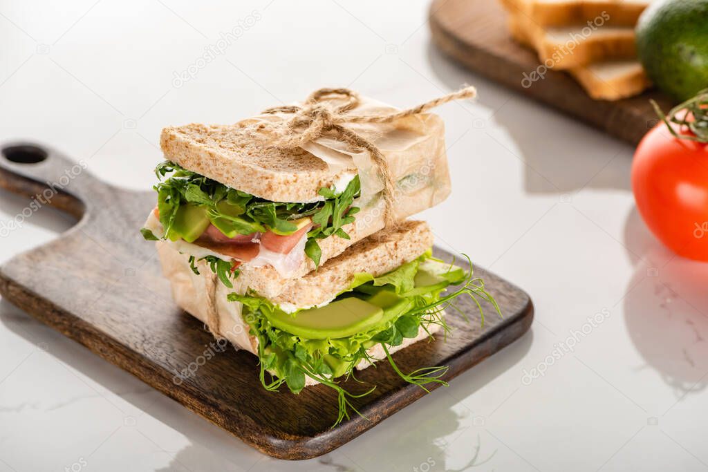 selective focus of fresh green sandwiches with avocado and meat on wooden cutting board on white marble surface