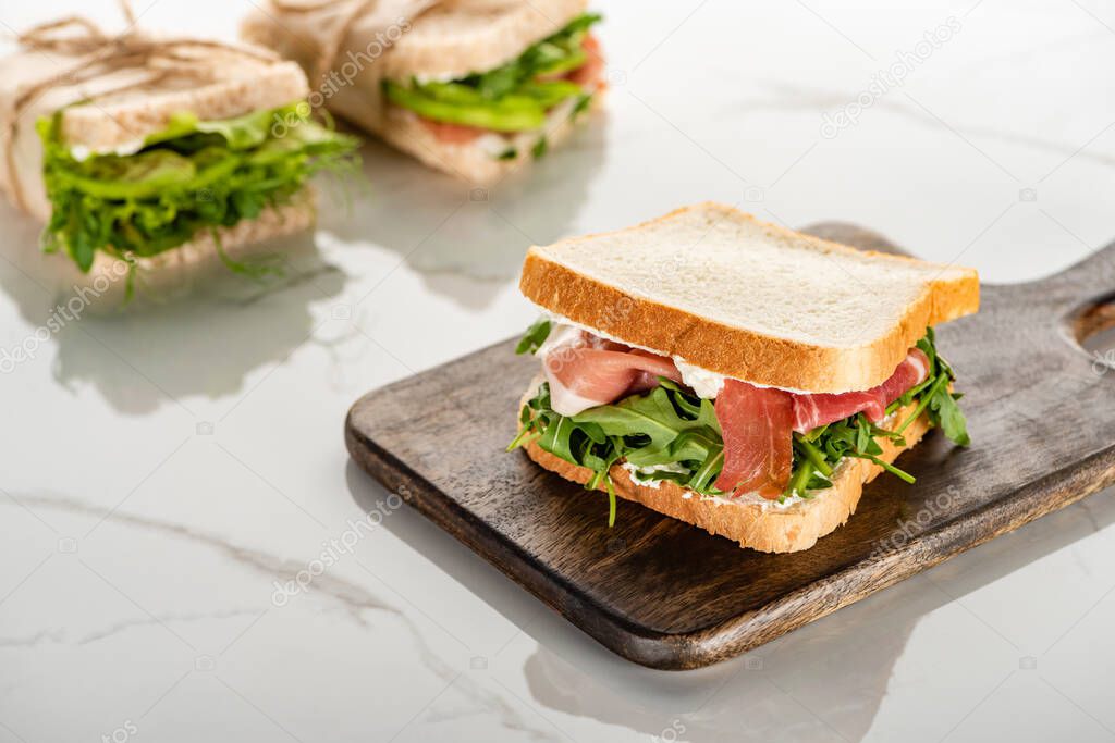 selective focus of fresh green sandwich with arugula and prosciutto on wooden cutting board on white marble surface