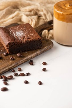 Kahve çekirdekleri, kahve ve beyaz arka planda kumaşla ahşap kesim tahtasındaki nefis browni parçası.