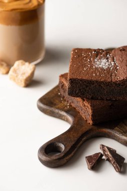 Tahta kesme tahtasındaki lezzetli browni parçalarının seçici odağı beyaz arka planda bardakta kahvenin yanında çikolata.
