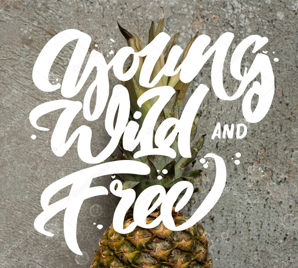 top view of ripe pineapple on grey concrete surface with young, wild and free illustration