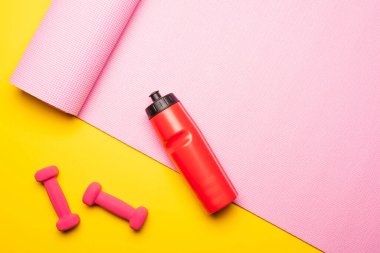 Kırmızı spor şişesinin üst görüntüsü, pembe spor paspası ve sarı arka planda dambıllar.