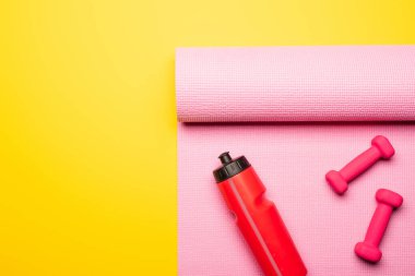 Kırmızı spor şişesinin üst görüntüsü, pembe spor paspası ve sarı arka planda dambıllar.