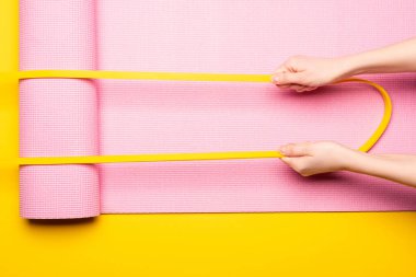 Kırpılmış görüntüde, sarı arka planda pembe fitness paspası üzerinde elastik bant tutan kadın görülüyor.