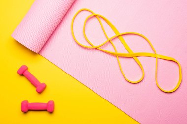 Sarı arka plandaki dambılların yanında pembe fitness minderinde direnç bandının üst görüntüsü