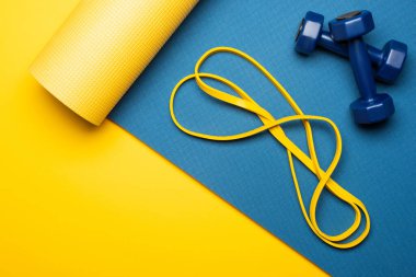 Sarı zemin üzerinde dambıllar ve direniş bandı olan mavi fitness paspasının üst görüntüsü