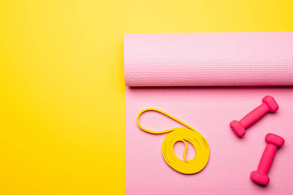 top view of pink dumbbells and elastic band on pink fitness mat on yellow background