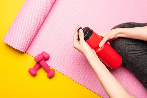在黄色背景的哑铃旁边 一个女人坐在粉色的健身垫上 一边拿着运动瓶子 一边看 — 图库照片