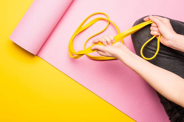 cropped view of woman sitting with resistance band on pink fitness mat on yellow background