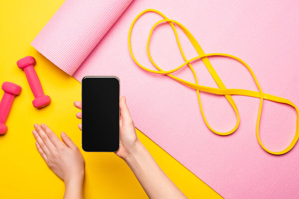 cropped view of woman holding smartphone near resistance band on pink fitness mat and dumbbells on yellow background