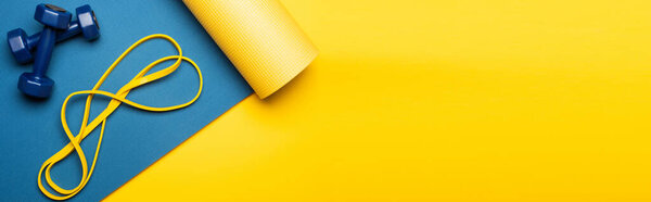 top view of blue fitness mat with dumbbells and resistance band on yellow background, panoramic shot