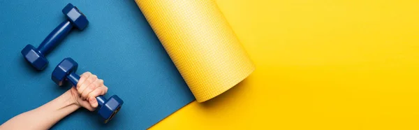 黄色の背景に青いフィットネスマットにダンベルを保持する女性のクロップドビュー パノラマショット — ストック写真