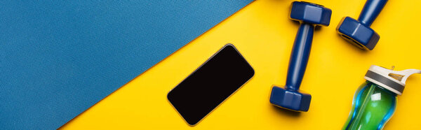 top view of blue fitness mat with dumbbells, smartphone and sports bottle on yellow background, panoramic shot