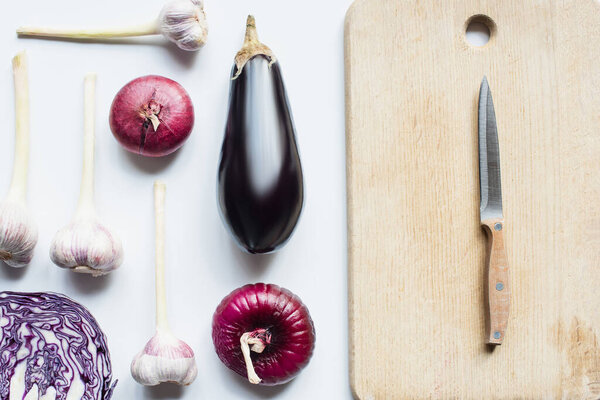 flat lay with red onion, red cabbage, eggplant and garlic near wooden cutting board with knife on white background