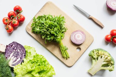 top view of ripe vegetables and knife near wooden cutting board with parsley and onion on white surface, panoramic shot clipart