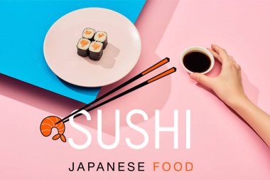 Elinde soya sosu tutan bir kadının somon balığı ve suşi Japon yemeğinin yanında mavi, pembe arka planda olduğu kesikli görüntüsü.
