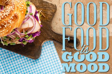 top view of fresh bagel with meat on wooden cutting board and plaid napkin near good food is good mood lettering  clipart