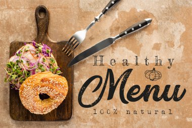 top view of bagel with meat on cutting board with cutlery near the healthy menu lettering on textured surface  clipart
