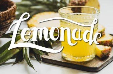 Bardaktaki taze ananas suyunun seçici odağı kesme tahtasındaki lezzetli meyvenin yanında ve beyaz harflerdeki limonata. 