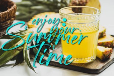 selective focus of pineapple juice in glass near delicious fruit on cutting board and enjoy your summer time lettering on white  clipart