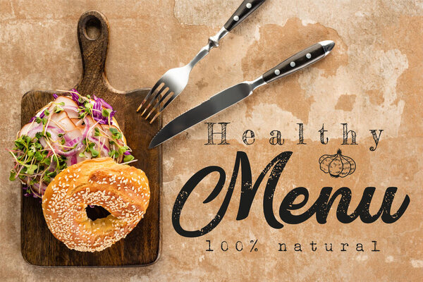 top view of bagel with meat on cutting board with cutlery near the healthy menu lettering on textured surface 