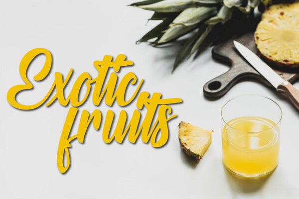 delicious pineapples on wooden cutting board with knife near glass of orange juice and exotic fruits lettering on white 