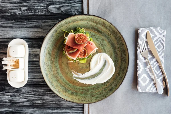 Vue de dessus du prosciutto gastronomique sur assiette et fourchette avec couteau sur table en bois — Photo de stock