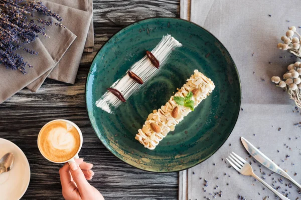 Abgeschnittene Aufnahme einer Person, die eine Tasse Cappuccino hält, während sie leckeres Dessert mit Mandeln isst — Stockfoto