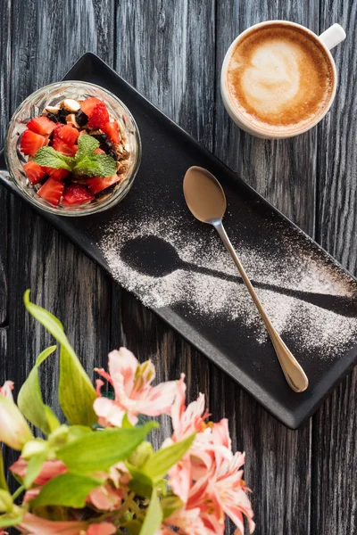 Vista superior de sabroso postre saludable con frutas, cuchara, taza de café y flores en la mesa de madera - foto de stock