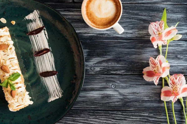 Blick von oben auf süßes, schmackhaftes Dessert auf Teller, Tasse Cappuccino und schöne Blumen auf Holztisch — Stockfoto