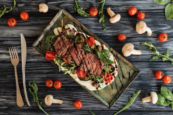 Vista superior de delicioso bistec asado, tenedor con cuchillo y verduras en mesa de madera - foto de stock