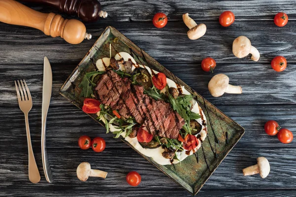 Blick von oben auf köstlich gekochtes Steak mit Gemüse, Gabel mit Messer und Gewürzen auf Holztisch — Stockfoto