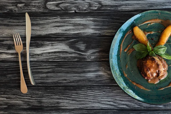 Viande gastronomique avec sauce et feuilles de basilic sur assiette, fourchette et couteau sur table en bois — Photo de stock