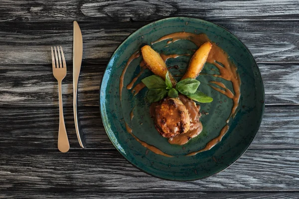 Вкусное приготовленное мясо с соусом и листьями базилика на тарелке, вилка и нож на деревянном столе — стоковое фото
