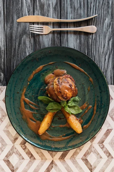 Delicious meat with sauce and basil leaves, fork and knife on wooden table — Stock Photo