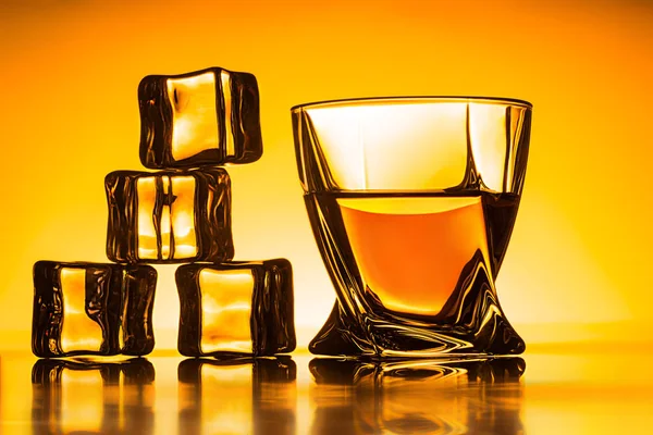 Glass with whiskey and ice cubes near on orange background — Stock Photo